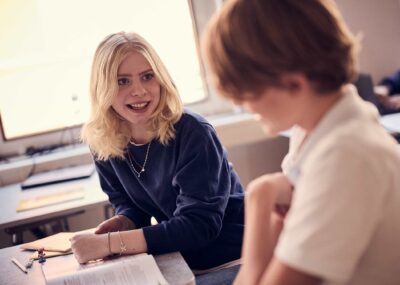 Två elever i ett klassrum på Snitz Grundskola sitter och pratar med varandra, de har läroböcker på bänken framför sig.