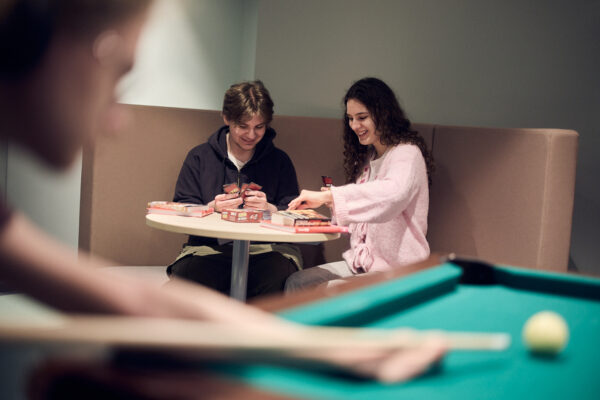 Två elever spelar kortspel tillsammans, medan andra elever spelar biljard framför.
